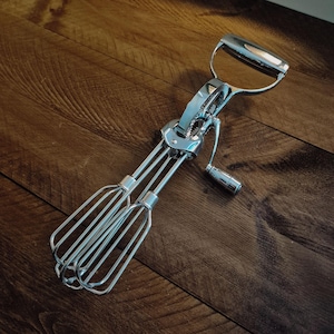 Könnte beinhalten: Ein verchromter Vintage-Handmixer mit Griff und Getriebemechanismus. Der Mixer hat zwei Sätze Drahtschläger und eine kleine Kurbel. Er liegt auf einer Holzoberfläche.