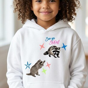 May include: White hooded sweatshirt featuring two raccoons and colorful stars. The text "See ya SOON!" is printed above the raccoons. The raccoons are in a playful pose, and the stars are pink, blue, and green.