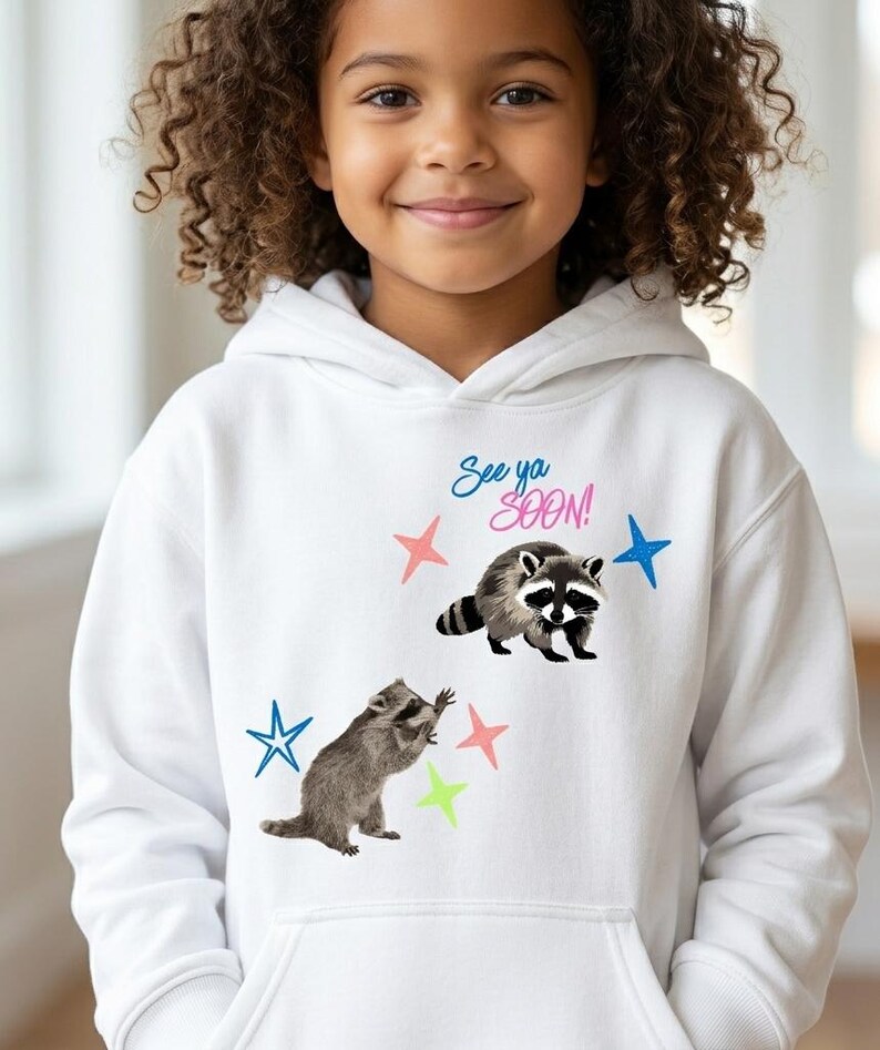 May include: White hooded sweatshirt featuring two raccoons and colorful stars. The text "See ya SOON!" is printed above the raccoons. The raccoons are in a playful pose, and the stars are pink, blue, and green.