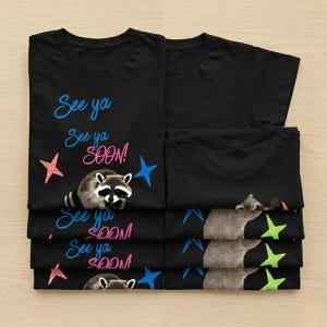 May include: Black t-shirts with a raccoon graphic and the text "See ya See ya SOON!" in blue and pink. The design includes pink and blue stars. The shirts are folded and stacked.