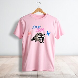 May include: A light pink t-shirt featuring a raccoon illustration with the text "See ya SOON!" above it. The design includes a pink and blue star on either side of the raccoon. The shirt is hanging on a wooden hanger.