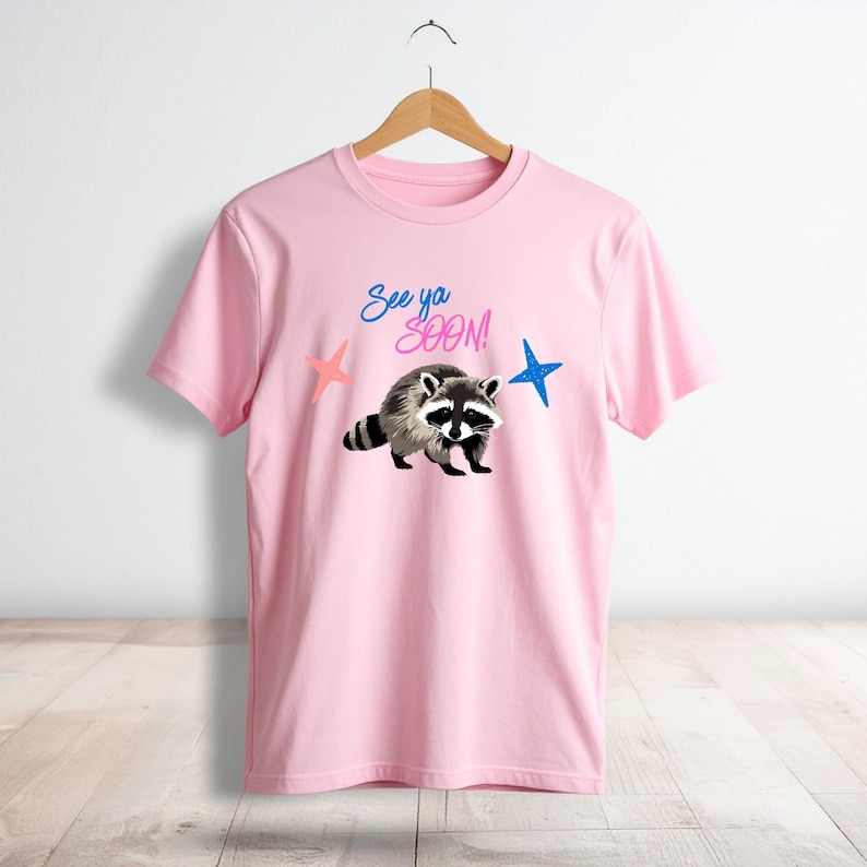 May include: A light pink t-shirt featuring a raccoon illustration with the text "See ya SOON!" above it. The design includes a pink and blue star on either side of the raccoon. The shirt is hanging on a wooden hanger.