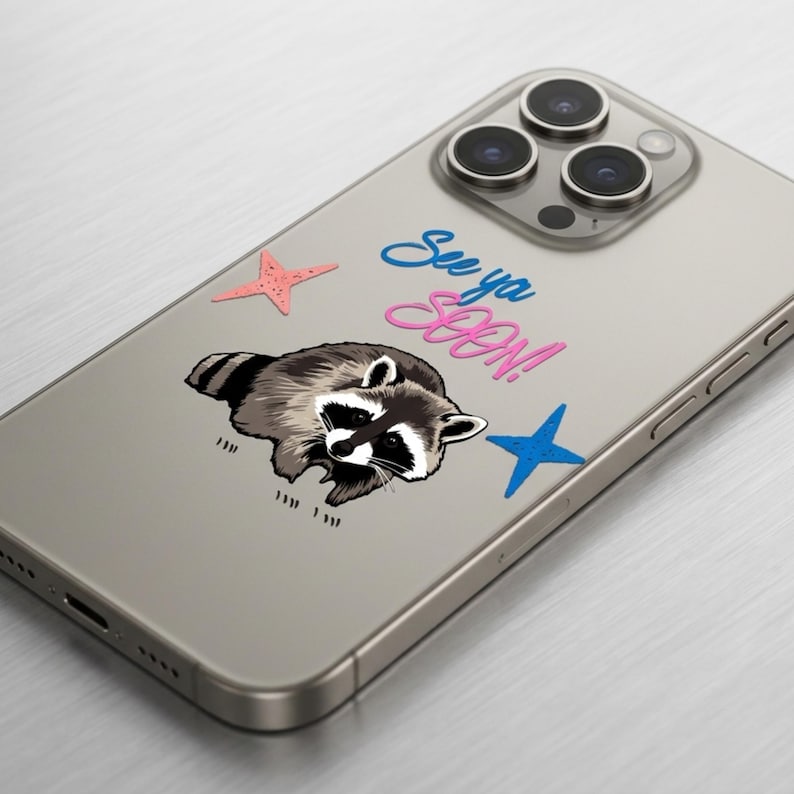 May include: A phone case featuring a raccoon illustration with the text "See ya SOON!" in blue and pink. The design includes a pink starfish and a blue star. The case is on a silver phone.