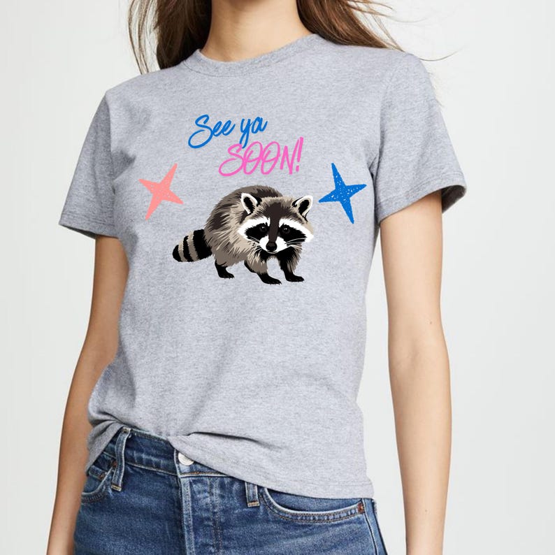 May include: Gray t-shirt featuring a raccoon illustration with the text "See ya SOON!" in blue and pink. The design includes pink and blue star accents. The shirt is a casual, everyday wear item.