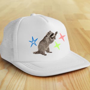 May include: White trucker hat featuring a raccoon illustration and colorful stars. The raccoon is gray and black, with a playful pose. The stars are blue, pink, and green. The hat has a mesh back.