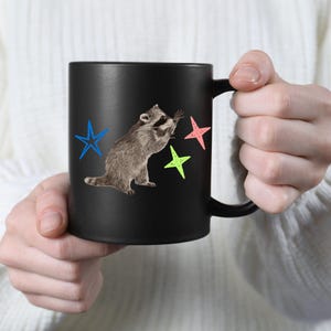 May include: Black ceramic mug featuring a raccoon reaching for colorful stars. The raccoon is depicted in shades of gray and brown, with blue, pink, and green star accents. The mug has a curved handle.
