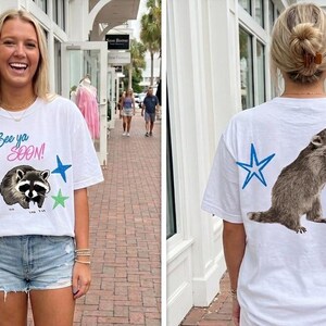 May include: White t-shirt with a raccoon graphic and the text "See ya SOON!" on the front. The back of the shirt features a raccoon and star graphics. The shirt is a casual, everyday wear item.