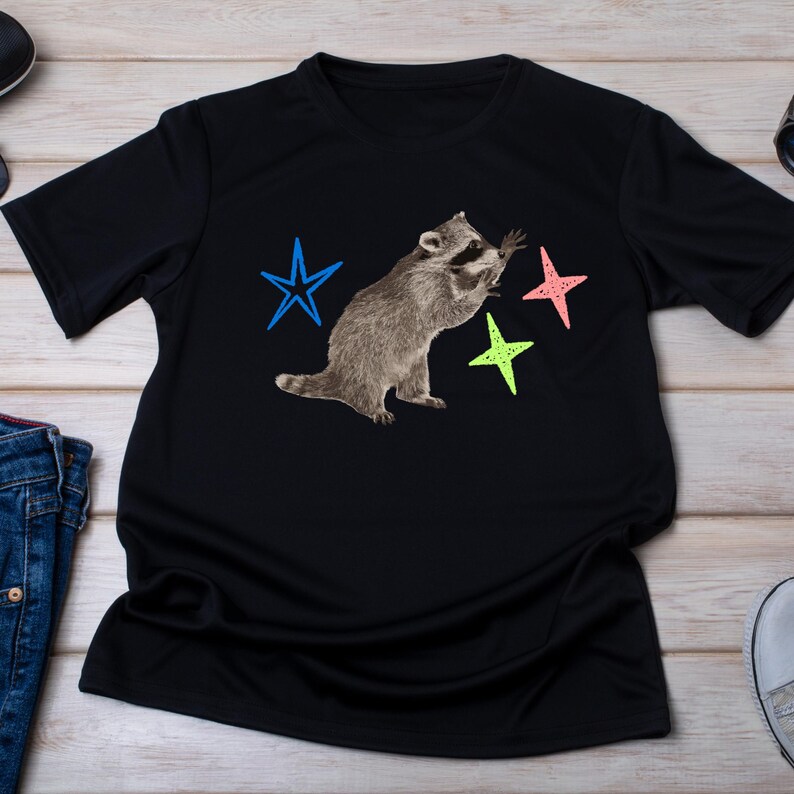 May include: Black t-shirt featuring a raccoon with outstretched paws and three colorful stars. The raccoon is depicted in shades of gray and brown. The stars are blue, pink, and green. The shirt is made of a soft material.