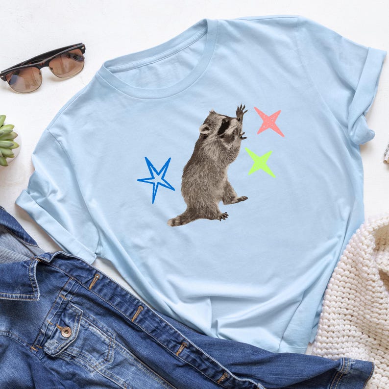 May include: Light blue t-shirt featuring a raccoon graphic reaching for colorful stars. The raccoon is in shades of brown and gray. The stars are blue, pink, and green. The shirt is made of a soft material.