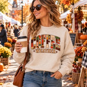 May include: A light heather grey crewneck sweatshirt featuring the text COFFEE WEATHER in a bold, block-letter font. The letters are filled with a multi-coloured patchwork pattern of autumn-themed prints, including plaid, leaves, and dots.