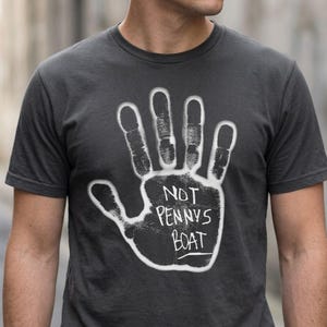 Può includere: T-shirt grigio scuro con un disegno di impronta di mano bianca e la scritta "NOT PENNYS BOAT". La grafica è centrata sul petto. La maglietta ha maniche corte e girocollo.