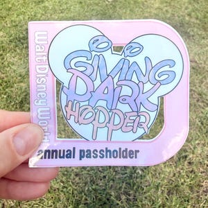 Può includere: Un adesivo rosa e blu con le parole "Swing Dark Hopper" in un design a orecchie di Topolino. L'adesivo riporta anche "Walt Disney World" e "annual passholder".
