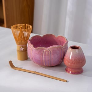 May include: A matcha tea set comprising a bamboo whisk, a scoop, a pink ceramic bowl with a scalloped edge, and a small pink ceramic tea bowl. The whisk has a flower decoration. The set is arranged on a white surface.