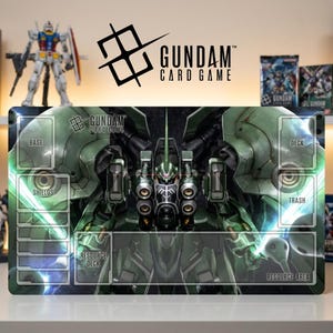 Puede incluir: Un tapete de juego rectangular de Gundam Card Game con una gran ilustración de un mobile suit verde. El tapete incluye zonas designadas etiquetadas como Base, Shields, Deck, Trash, Resource Deck y Resource Area, con el logo de Gundam Card Game en la esquina superior izquierda.