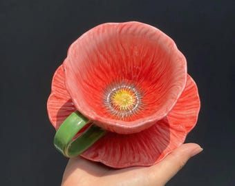 May include: A ceramic teacup and saucer set designed to resemble a poppy flower. The cup and saucer are red with a yellow and grey centre. The handle is green.