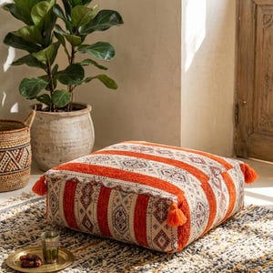 Op de afbeelding: Een rechthoekige vloerkussen met een oranje en crèmekleurig streepjespatroon en decoratieve kwastjes. De kussen heeft een geometrisch ontwerp en is geplaatst op een patroonkleed. Een plant in pot en een gevlochten mand staan op de achtergrond.