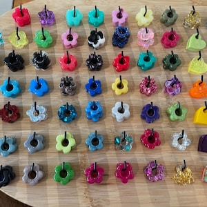 Peut inclure: Une collection de breloques colorées en forme de fleurs avec des boucles noires. Les breloques sont de différentes couleurs, dont le rouge, le violet, le vert et le bleu, et sont disposées sur une surface en bois. Certaines breloques ont des paillettes.