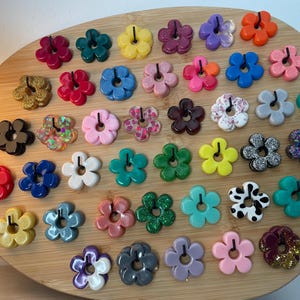 Peut inclure: Une collection d'accessoires en forme de fleurs colorées, chacune avec un petit trou au centre. Les fleurs sont de différentes couleurs, notamment rouge, bleu, jaune et rose, et certaines ont des détails pailletés ou à motifs. Elles sont disposées sur une surface en bois.