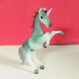 May include: A glass figurine of a rearing unicorn. The unicorn is primarily gray with a white underbelly and legs. The mane and tail are a reddish-purple, and the horn is pink. The figurine is on a white surface with a red background.
