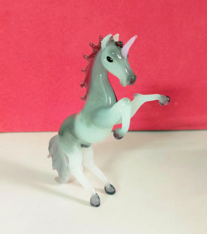 May include: A glass figurine of a rearing unicorn. The unicorn is primarily gray with a white underbelly and legs. The mane and tail are a reddish-purple, and the horn is pink. The figurine is on a white surface with a red background.