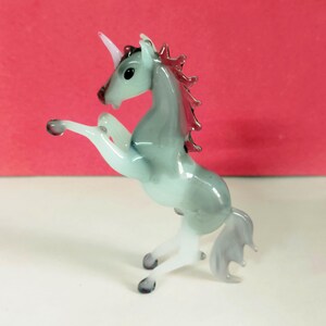 May include: A glass figurine of a rearing unicorn. The unicorn has a pale blue body, white legs, and a pink horn and mane. The figurine is on a white surface with a red background.