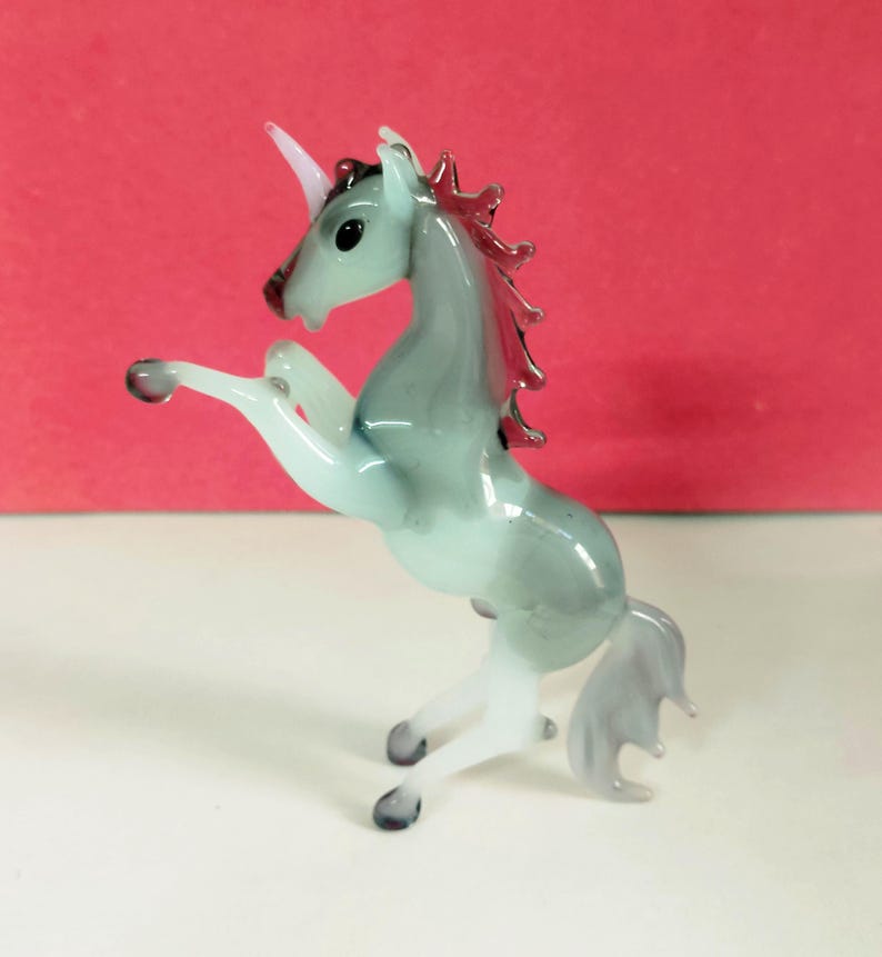 May include: A glass figurine of a rearing unicorn. The unicorn has a pale blue body, white legs, and a pink horn and mane. The figurine is on a white surface with a red background.