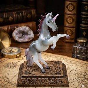 May include: A glass unicorn figurine in shades of gray, white, and purple, rearing up on its hind legs. The unicorn has a pink horn and mane, and is mounted on a wooden base with carved details. The figurine is displayed on a map.