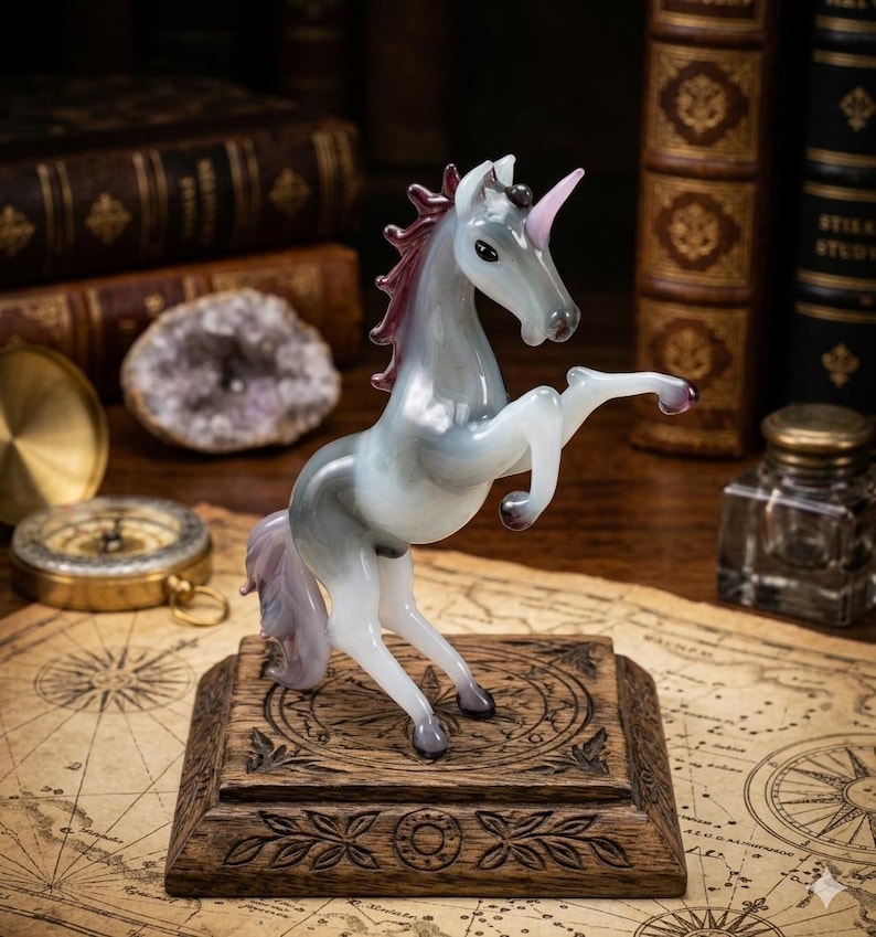 May include: A glass unicorn figurine in shades of gray, white, and purple, rearing up on its hind legs. The unicorn has a pink horn and mane, and is mounted on a wooden base with carved details. The figurine is displayed on a map.