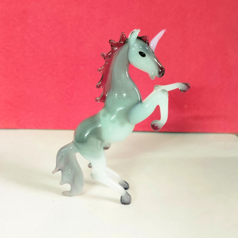 May include: A glass unicorn figurine in a rearing pose. The unicorn has a white horn, a gray and light blue body, and a red mane. The figurine is on a white surface with a red background.