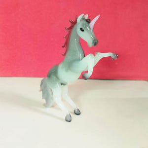 May include: A glass unicorn figurine in a rearing pose. The unicorn has a gray body with a white mane and tail. The horn is pink, and the mane is a darker red. The figurine is on a white surface with a red background.