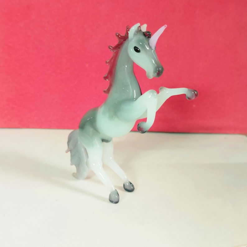 May include: A glass unicorn figurine in a rearing pose. The unicorn has a gray body with a white mane and tail. The horn is pink, and the mane is a darker red. The figurine is on a white surface with a red background.