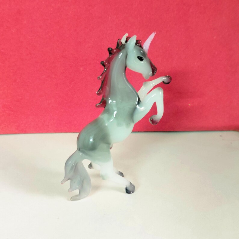 May include: A glass figurine of a rearing unicorn. The unicorn is primarily gray with white legs and a pink horn. The mane and tail have dark accents. The figurine is on a white surface with a red background.