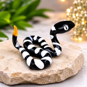 May include: A glass snake figurine featuring a glossy black body with irregular white bands and a bright orange tail tip. The snake is coiled in a resting position with its head raised, showcasing white oval eyes on a smooth, reflective surface.