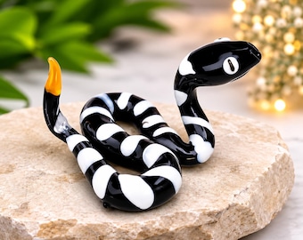 May include: A glass snake figurine featuring a glossy black body with irregular white bands and a bright orange tail tip. The snake is coiled in a resting position with its head raised, showcasing white oval eyes on a smooth, reflective surface.