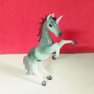 May include: A glass unicorn figurine in shades of gray, white, and pink. The unicorn is rearing up on its hind legs, with a pink horn and a flowing mane. The figurine has a glossy finish and is set against a red background.