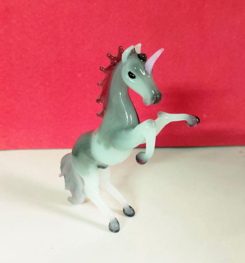 May include: A glass unicorn figurine in shades of gray, white, and pink. The unicorn is rearing up on its hind legs, with a pink horn and a flowing mane. The figurine has a glossy finish and is set against a red background.