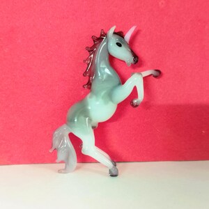 May include: A glass unicorn figurine in a rearing pose. The unicorn has a white horn, a gray and white body, and a dark red mane. The figurine is on a white surface with a red background.