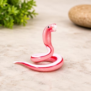 Op de afbeelding: Een klein glazen beeldje van een opgerolde slang in roze en witte tinten. De slang heeft een opgeheven kop, een gladde glanzende afwerking, twee kleine witte giftanden en piepkleine zwarte oogjes. Het lichaam is doorschijnend roze van kleur.