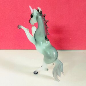 May include: A glass unicorn figurine in a rearing pose. The unicorn has a white horn, a gray and white body, and a flowing tail. The figurine is on a white surface with a red background.