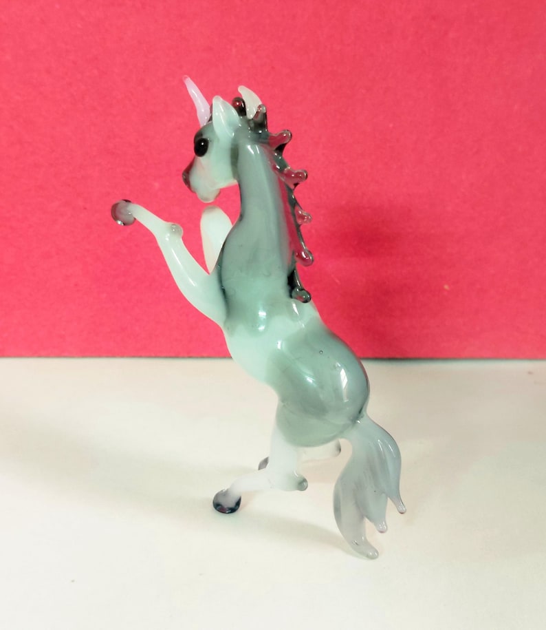 May include: A glass unicorn figurine in a rearing pose. The unicorn has a white horn, a gray and white body, and a flowing tail. The figurine is on a white surface with a red background.