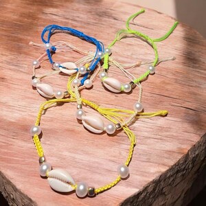 May include: Several handmade bracelets with natural seashells, faux pearls, and colourful cords. The bracelets are in blue, yellow, and green, with a rustic, beachy style. Each bracelet has a unique design, perfect for summer.