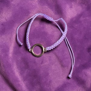 Peut inclure: Un bracelet macramé violet clair avec une pièce maîtresse circulaire de couleur or. Le bracelet est posé sur un fond en tissu violet. Le bracelet a des nœuds coulissants réglables.