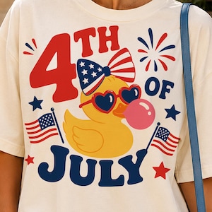 May include: An off-white short-sleeve t-shirt featuring a graphic of a yellow rubber duck wearing heart-shaped sunglasses and a patriotic bow. The text reads 4th of July, accompanied by American flag illustrations, stars, and fireworks.