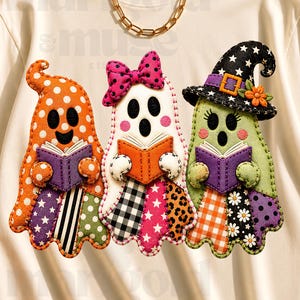 May include: A cream-colored short-sleeve t-shirt featuring a graphic of three ghosts reading books. The ghosts are decorated with various patterns including polka dots, plaid, and stars. One ghost wears a pink bow, and another wears a witch hat.