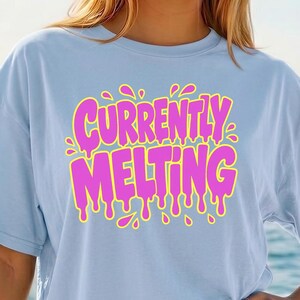 May include: Light blue t-shirt with the text "CURRENTLY MELTING" in pink with yellow outlines and dripping details. The shirt is a casual style, suitable for everyday wear.