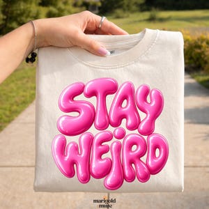 Peut inclure: T-shirt crème avec l'inscription "STAY WEIRD" en lettres roses à bulles. La main tient le t-shirt. Le logo "marigold muse" est visible en bas à droite.