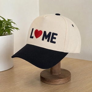 Puede incluir: Una gorra de béisbol de color crema con visera negra y un botón oscuro en la parte superior. El frente presenta letras azul marino bordadas que forman L-M-E con un corazón rojo brillante reemplazando la O, creando el texto L-corazón-ME.