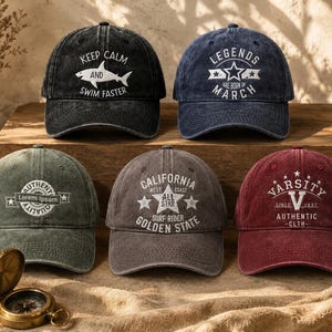 Puede incluir: Cinco gorras de béisbol en negro, azul marino, verde oliva, gris carbón y granate, cada una con textos y gráficos bordados en blanco, incluyendo 'Keep Calm and Swim Faster', 'Legends are Born in March', 'California' y 'Varsity'.