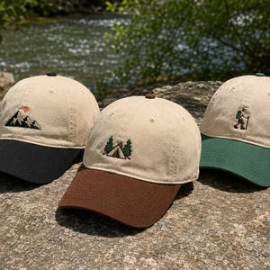 Puede incluir: Tres gorras de béisbol con coronas beige y viseras en contraste. La gorra de la izquierda tiene visera negra y bordado de montaña. La central tiene visera marrón y bordado de tienda de campaña. La de la derecha tiene visera verde y bordado de excursionista.