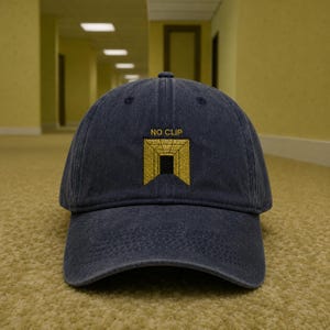 Puede incluir: Una gorra de béisbol azul marino con un diseño bordado de una forma geométrica amarilla en forma de túnel con un centro negro y el texto NO CLIP colocado directamente sobre el gráfico en una fuente amarilla simple sin remates.